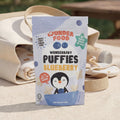 Wunderbaby Puffies Blueberry packaging on a wooden surface with a blanket and basket in the background.